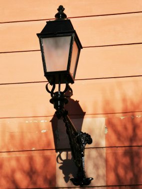 The classic black street lamp is affixed to a peach-colored wall, creating a charming atmosphere. The afternoon sunlight accentuates its elegant design and casts distinct shadows. clipart