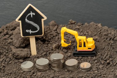 Dünyanın arka planında gümüş paralarla sarı bir kazıcının oyuncak modeli. Eğik ya da maliyet inşa kavramı