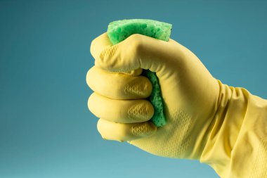 Hand in yellow rubber glove holds sponge for washing dishes and cleaning, hand clenches sponge into fist on blue background