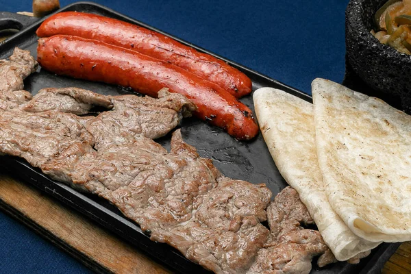 Meksika usulü biftek ve domuz eti, yanında iki quesadilla ve molcajete sosu, ahşap bir tahta üzerinde ve mavi bir masada bir matal comal..