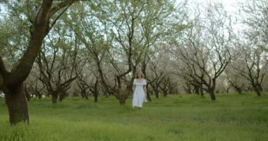 Zarif bir kadın, dalgalı beyaz bir elbise giymiş, huzurlu ağaçların arasında neşeyle yürür.