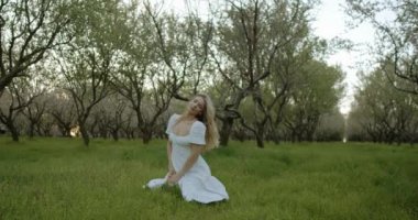 Huzurlu bir sahnede akıcı beyaz elbiseli bir kadın, büyüleyici bir meyve bahçesinde yemyeşil ağaçlar içinde.