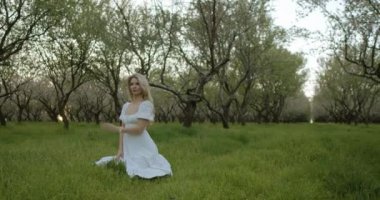 Akan beyaz elbiseli güzel bir kadın, uzun ağaçlarla çevrili canlı yeşil bir tarlada zarif bir şekilde diz çöker.