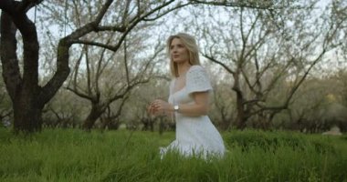 Akan beyaz elbiseli genç bir kadın çiçek açan ağaçlarla dolu yemyeşil bir tarlada güzelliği takdir eder.
