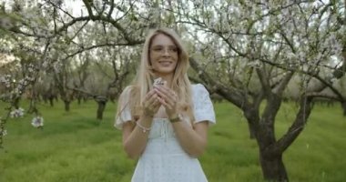 Beyaz elbiseli neşeli bir kadın çiçek açan bir bahçede çiçeklere hayran, baharın güzelliğini yakalıyor..