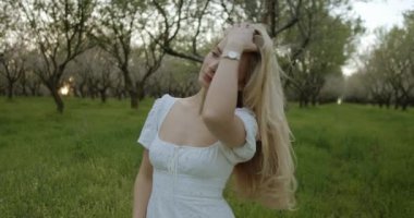 Akan beyaz bir elbiseyle süslenmiş zarif bir figür, sakin ve resimsel bir meyve bahçesinin arka planına kurulur.