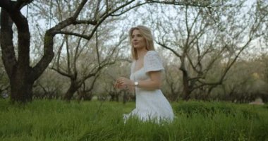 Akan beyaz elbiseli genç bir kadın çiçek açan ağaçlarla dolu yemyeşil bir tarlada güzelliği takdir eder.