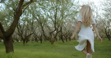 Dalgalı beyaz elbiseli genç bir kadının renkli çiçeklerin açtığı bir meyve bahçesinde gezindiği huzurlu bir sahne.