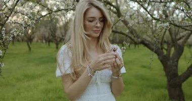 Huzurlu ve sakin bir sahne, çiçek açan ağaçların arasında dalgalı beyaz bir elbise giyen güzel bir kadını gösteriyor.