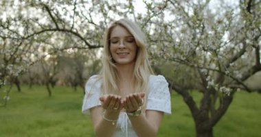 Çiçek açan bir meyve bahçesinde ellerini zevkle açık tutan dalgalı beyaz elbiseli bir kadının sakin bir sahnesi.