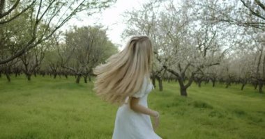 Beyaz elbiseli neşeli bir kadın hayat ve renk dolu bir meyve bahçesinde canlı bir bahar gününün tadını çıkarıyor.