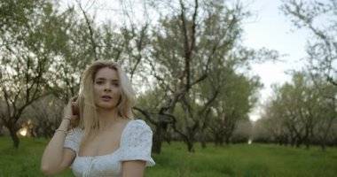 Huzurlu bir sahnede, dalgalı beyaz elbiseli bir kadın serpilen ağaçların arasında zarif bir şekilde dikiliyor.