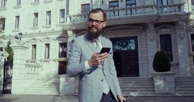 Zarif bir şekilde giyinmiş modern bir işadamı telefonuna dikkat çekmeden göz kamaştırıcı bir binanın önünde bakıyor.