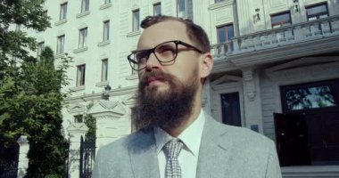 Şık giyinmiş, gözlüklü ve bakımlı sakallı bir adam tarihi bir binanın önünde duruyor.