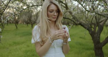 Huzurlu ve sakin bir sahne, çiçek açan ağaçların arasında dalgalı beyaz bir elbise giyen güzel bir kadını gösteriyor.