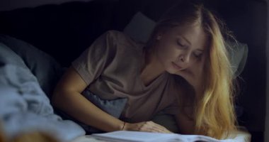 Düşüncelere dalmış genç bir kadın, rahat yatağında büyüleyici bir kitap okuyor. Nazik, sıcak bir ışıkla aydınlatılıyor.