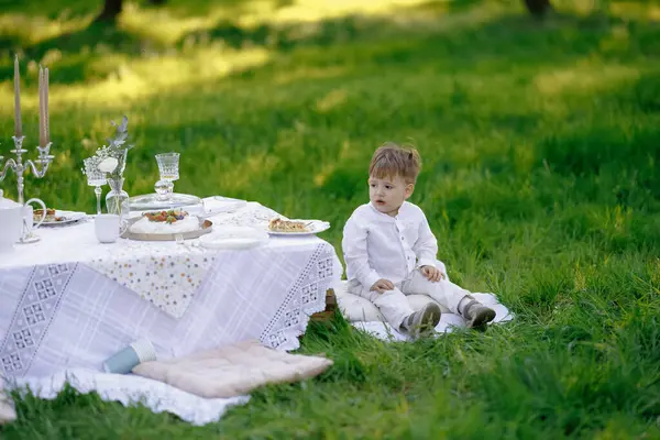 Küçük bir çocuk piknik masasının önüne konmuş renkli bir battaniyenin üzerinde rahatça oturuyor ve güzel bir günün tadını çıkarıyor.
