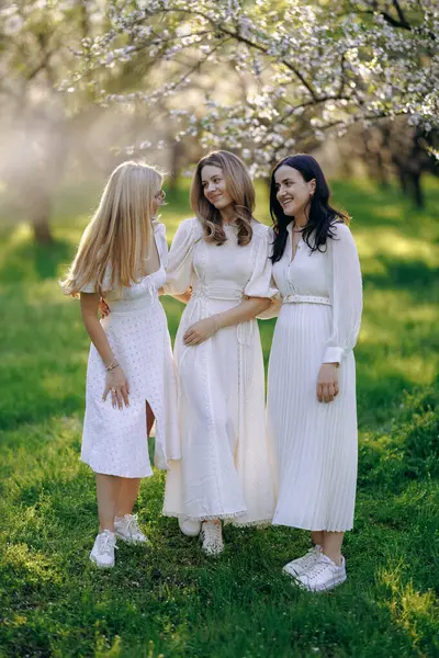 Üç kadın, hepsi de güzel beyaz elbiseler giymiş güzel bir park ortamında yan yana durmuş birlikte vakit geçiriyorlar.