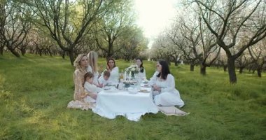 Çiçek açan bir meyve bahçesinde huzurlu ve huzurlu bir toplantı gerçekleşiyor. Hafif elbiseler ve doğal güzellikler sergileniyor.