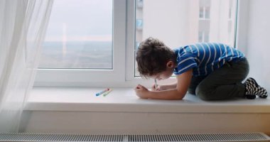 Bu büyüleyici videoda, genç bir çocuk pencere pervazında oturmuş, yaratıcılığını teknoloji, boya kalemleri ve kalemlerle ifade etmek için sanat malzemelerini kullanıyor.