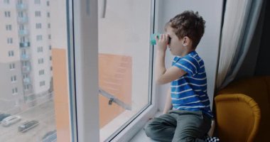 Pencere pervazına tünemiş genç bir çocuk dürbünle dünyayı keşfediyor, gözleri ışıl ışıl parlıyor, çocukluk masumiyetini ve merakını somutlaştırıyor.