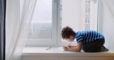 Genç bir çocuk pencerenin pervazında oturup dışarıdaki dünyadan esinlenerek renkli kalemler, kalemler ve pastel kalemlerle yaratıcılığını ifade ediyor.