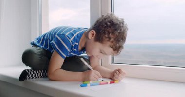 Genç bir çocuk mutlu bir şekilde pencere eşiğinde oturmuş renkli kalemlerle çizmeye odaklanmış yaratıcılığını ve hayal gücünü sergiliyor.
