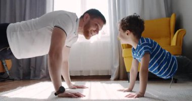 Bir adam ve genç bir çocuk eğlenceli bir fitness aktivitesinin keyfini çıkarıyorlar, rahat bir oturma odasında şınav çekiyorlar, kahkahalarını paylaşıyorlar ve bağlarını gösteriyorlar.