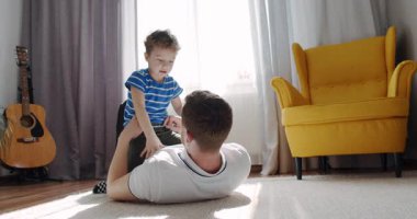 Bir erkek ve genç bir çocuğun kahkaha ve neşeyle dolu sıcak bir salonun zemininde oynadığı iç açıcı bir sahne gözler önüne seriliyor