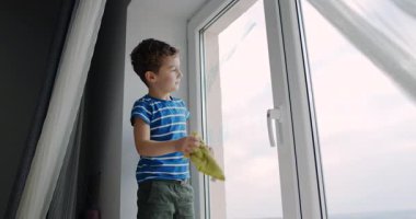 Genç bir çocuk bir bez ve süngerle bir pencereyi özenle temizler. Daha sonra, dışarı bakar, dünyaya olan merakını yansıtır.