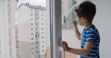 Genç bir çocuk, bir keçeli kalemle pencereye çizerek yaratıcılık gösteriyor. Dışarıya bakıyor, merakını paylaşıyor, sonra pencereyi temizliyor, keşifteki masumiyet ve neşeyi yansıtıyor.