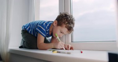 Genç bir çocuk, bir pencere eşiğinde, çizim ve yazma yoluyla yaratıcılığını ifade ederek, kalemler ve bir defter kullanarak teselli buluyor. Bu sahne çocukluk masumiyetini ve neşesini yansıtır.