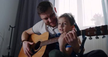 Sıcak bir oturma odasında, bir baba küçük oğluna gitar çalmayı öğretir, kahkaha ve müzikle dolu neşeli anları paylaşır.
