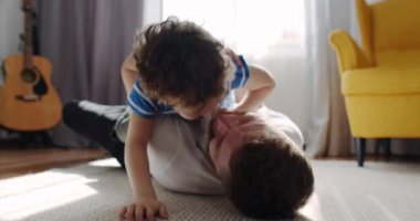 Sıcak bir oturma odasında, bir baba ve oğul neşe dolu bir anı paylaşıyor, yerde oynarken kahkahalarla kaynaşıyorlar, sevgi gösterisi yapıyorlar.