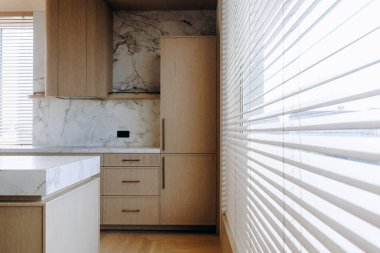 Zarif ahşap dolaplar, ferah bir buzdolabı, fonksiyonel bir lavabo ve şık jaluzilerle süslenmiş bir pencere içeren güzel tasarlanmış bir mutfak.