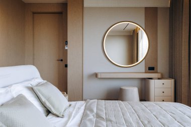 Konforlu bir yatak odasında, yumuşak döşemelerle süslenmiş rahat bir yatak ve duvarda zarif bir şekilde asılı yuvarlak ayna var.