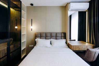 Rahat bir yatak odası, şık bir sandalye, ahşap bir şifonyer ve parlak bir pencere.