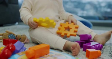 Küçük bir bebek yerde mutlu bir şekilde oturuyor, etraflarına çeşitli renkli oyuncaklar serpiştirilmiş bir oyun zamanı ile meşgul.