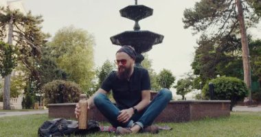 Sakallı bir adam sakin bir park gününün tadını çıkarır. Çimlerin üzerinde çeşmenin kenarında oturur, birasını yudumlar, sandviçin tadını çıkarır ve enstrüman çalar.