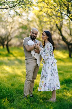 Güzel bir parkta, bir erkek ve bir kadın, doğanın canlı renkleri ve çiçek açan çiçekleriyle çevrili küçük bir bebeği sevgiyle kucaklıyorlar.