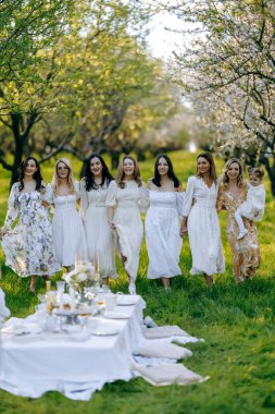 Bir grup zarif giyinmiş beyaz elbiseli kadın masanın etrafındaki güzel bir parkta yan yana duruyorlar.