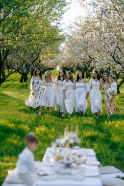 Beyaz elbiseler içinde zarif giyinmiş kadınların bir araya gelmesi bahar zamanı güzel bir masa etrafında dikiliyor.