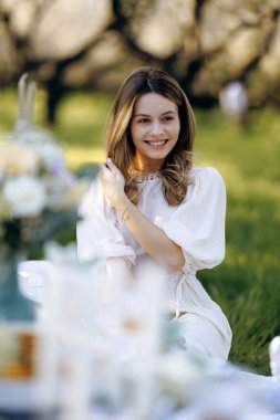 Zarif, beyaz bir elbise giymiş bir kadın hoş bir masanın önündeki yumuşak çimlerin üzerinde nazikçe oturuyor.