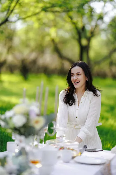 Güzel beyaz bir elbise giyen bir kadın zarif bir masada, doğanın sıcaklığı ve neşe ile çevrili bir şekilde huzur içinde oturuyor.