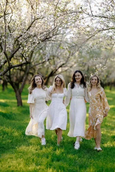 Göz kamaştırıcı beyaz elbiseler giymiş bir grup güzel kadın canlı bir parkta birlikte mutlu bir şekilde yürüyorlar.
