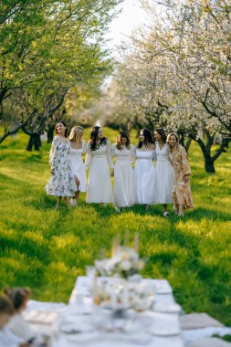 Zarif beyaz elbiseler giymiş bir grup kadın, güzel bir şekilde yerleştirilmiş bir masanın etrafında dikiliyorlar.