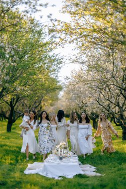 Zarif beyaz elbiseler giyen hoş bir grup kadın, canlı bir parkta güzel bir şekilde düzenlenmiş bir masanın etrafında neşeyle duruyorlar.