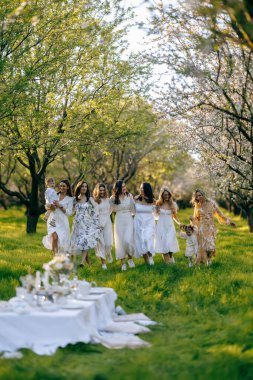 Zarif beyaz elbiseler giymiş, resim gibi bir grup kadın, yemyeşil bir alanda el ele tutuşup el ele tutuşurken görülebilir.