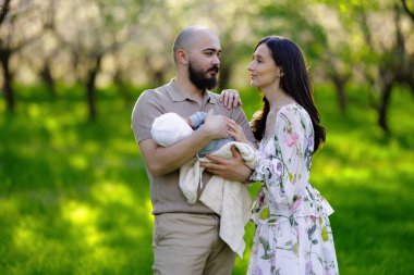 Güzel bir parkta, bir erkek ve bir kadın sevgi dolu bir bebeği kucaklıyorlar, huzurlu atmosferin ve birbirlerine eşlik etmenin tadını çıkarıyorlar.