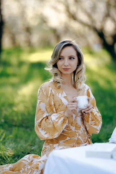 Güzel bir ortamda, canlı sarı bir elbise giymiş bir kadın, elinde sıcak bir fincan kahve tutarken yemyeşil çimlerin üzerinde rahatça oturuyor.